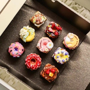 May include: A collection of miniature, decorative donuts in a dark box. The donuts feature various toppings, including sprinkles, flowers, and fruit. One donut has the text "Sprinkled with Love", and another says "Happy Birthday".