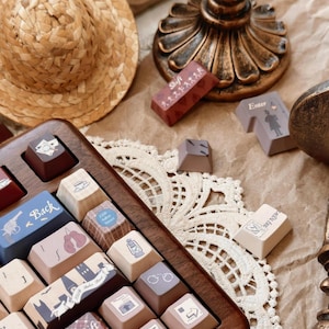 May include: A wooden keyboard with custom keycaps featuring illustrations of a city skyline, a detective, a magnifying glass, a pair of handcuffs, a boxing glove, a gun, a lantern, and other vintage-inspired designs. The keyboard is surrounded by decorative items, including a straw hat, a gold candlestick, and a lace doily.
