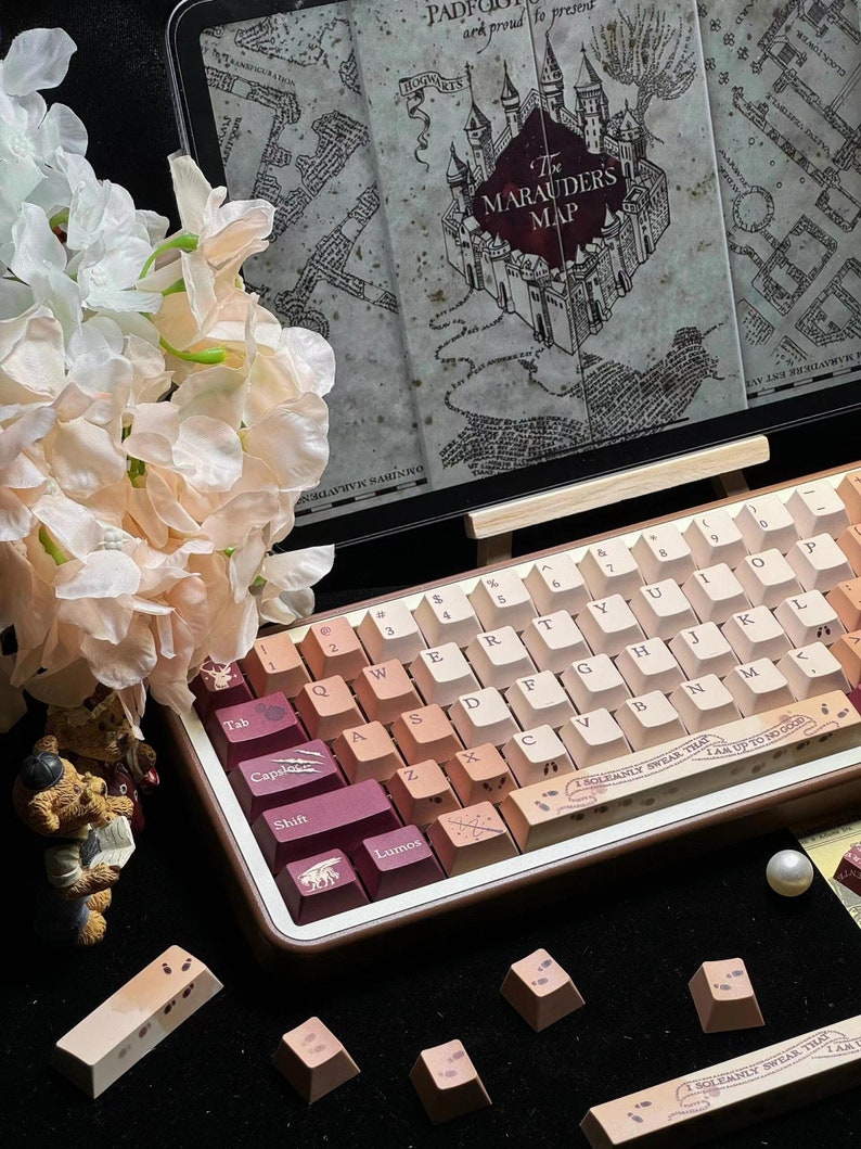 May include: A custom mechanical keyboard with gradient keycaps in shades of pink and beige. The keyboard features themed keycaps with text and symbols. A display behind the keyboard shows a map with the text "The Marauders Map".