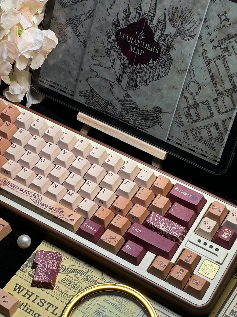 May include: A custom mechanical keyboard with a brown and cream color scheme. The keycaps feature various symbols and text, including "Lumos" and "Shift." The keyboard is placed in front of a tablet displaying "The Marauders Map."