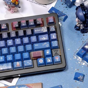 May include: A mechanical keyboard with blue and pink keycaps. The keycaps feature various illustrations and text, including "Elle" and "Know that my future is not just a dream." Decorative figurines and a lit candle are also present.