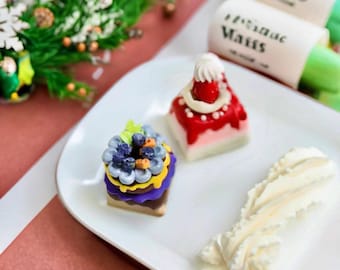 Könnte beinhalten: Ein weißer Teller mit Miniaturdesserts, darunter ein Kuchen mit Blaubeer- und gelbem Blumendesign, ein Kuchen mit roter Spitze und eine Spirale aus weißem Zuckerguss. Der Hintergrund umfasst einen Zweig mit Grün und zwei Rollen mit Text.