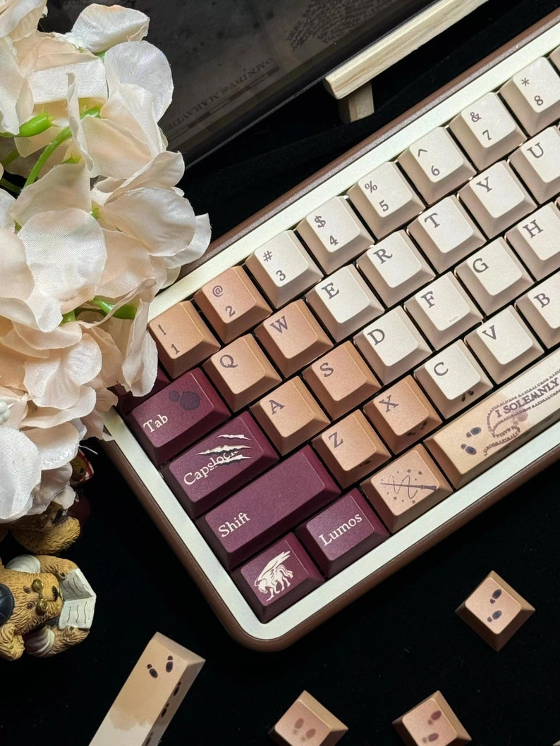 May include: A custom mechanical keyboard with a brown wooden frame and cream-colored keycaps. The keycaps feature a gradient of colors, including burgundy and tan. Some keys have themed icons and text, such as "Lumos" and "I SOLEMNLY".