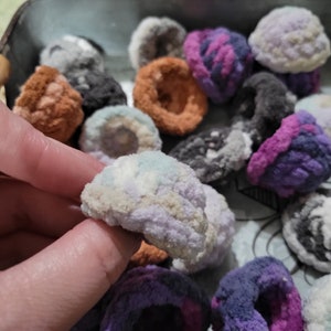 May include: A collection of colorful, fuzzy, crocheted, flower-shaped ornaments in a variety of colors, including purple, pink, blue, white, and gray.
