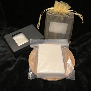 May include: A rectangular white bar of soap sealed in a clear plastic bag, accompanied by a black cardboard box with a rectangular cutout and a gold-colored sheer organza drawstring pouch, all arranged together on a dark background.
