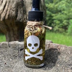 May include: A clear glass dropper bottle filled with dried herbs and botanical matter. The bottle features a white skull and crossbones decal on the front, a black dropper cap, and a bronze-toned pentagram charm tied with twine around the neck.