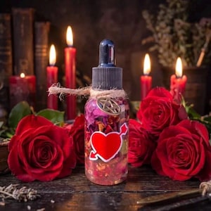 May include: A clear glass dropper bottle filled with liquid and dried flower petals, featuring a red heart sticker with an arrow and a small metal pentagram charm tied around the neck with twine, surrounded by several vibrant red roses.