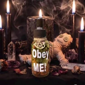May include: A clear glass dropper bottle filled with dried herbs and liquid, featuring a gold pentagram charm tied with twine around the neck. The bottle has the white text Obey ME! printed on the front, surrounded by black candles and a burlap doll.