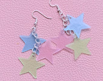 May include: A pair of drop earrings featuring multiple translucent star-shaped charms in pastel shades of blue, pink, and yellow. The stars are attached to silver-toned metal chains and ear hooks, creating a layered, multi-star design.