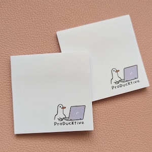 May include: Two square white sticky notes featuring a cartoon illustration of a white duck with an orange beak sitting at a light purple laptop. Below the illustration, the text ProDucktive is printed in a simple black sans-serif font.