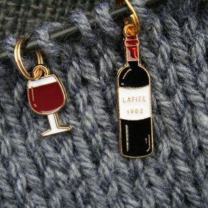 Könnte beinhalten: Zwei goldfarbene Maschenmarkierer mit Emaille-Anhängern. Ein Anhänger ist ein rotes Weinglas, gefüllt mit Rotwein. Der andere Anhänger ist eine schwarze Weinflasche mit einem goldenen Etikett, auf dem "Lafite 1982" steht.