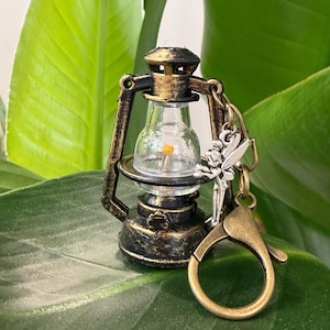 May include: Miniature antique-style lantern keychain with a clear glass globe and a warm, flickering LED light. The lantern is bronze-colored with a silver fairy charm and a gold-toned clasp. The keychain is set against a backdrop of green leaves.