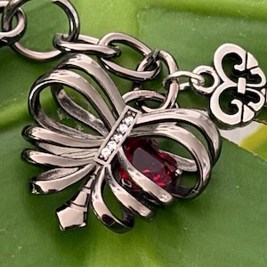 May include: A silver-toned charm bracelet with a bow-shaped pendant. The pendant features a red gemstone and small clear stones. The charm is attached to a chain with a decorative heart-shaped element.