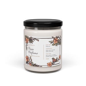 May include: A glass jar candle with a white label that reads "Pixie Perfume Fairy Garden Collection". The label has a floral design with orange and brown flowers. The candle is vegan and made with 100% natural soy wax and a 100% cotton wick. The burn time is 50-60 hours.