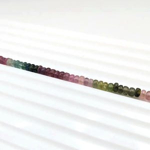 May include: A strand of round, multi-colored gemstone beads. The beads are pink, green, blue, and purple. The beads are arranged in a gradient pattern, starting with pink and ending with green.