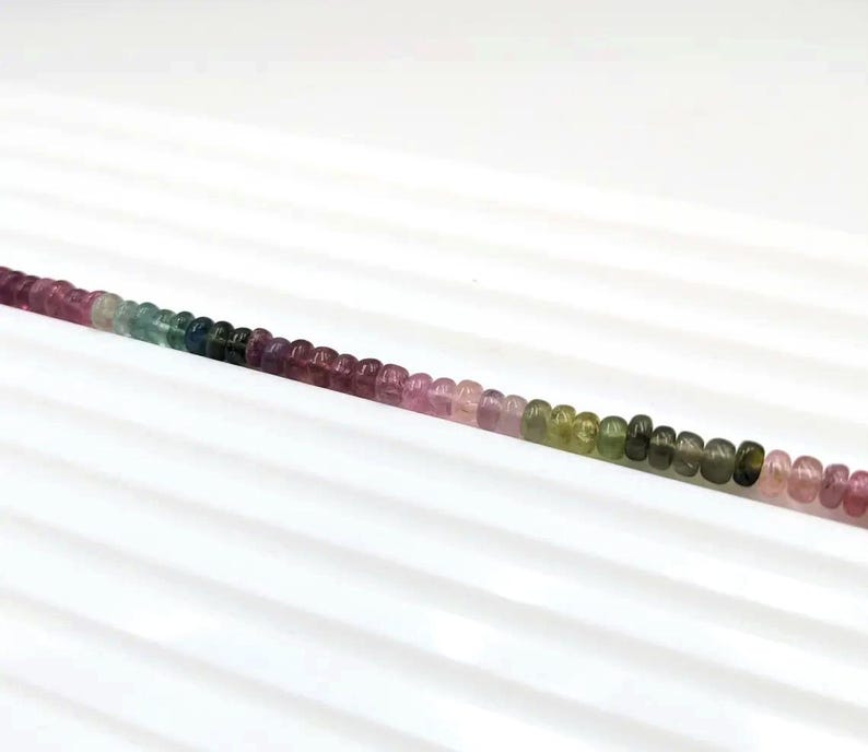 May include: A strand of round, multi-colored gemstone beads. The beads are pink, green, blue, and purple. The beads are arranged in a gradient pattern, starting with pink and ending with green.