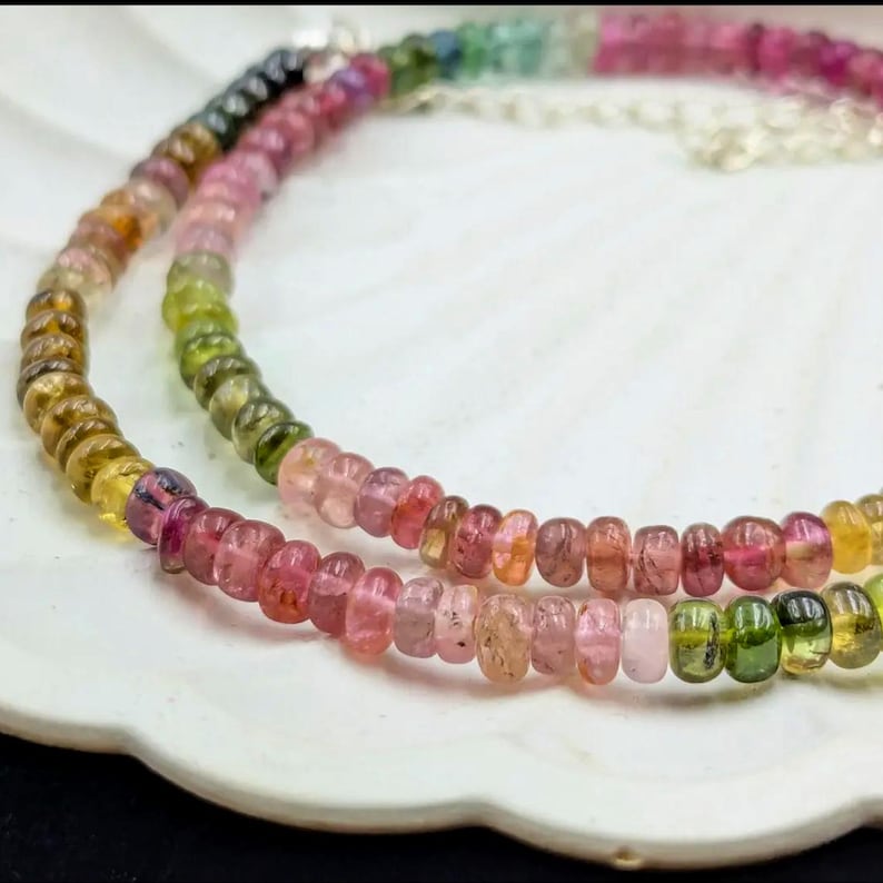 May include: A double strand necklace made of small, round, multi-colored gemstones. The necklace features a gradient of colors, starting with green and yellow, transitioning to pink and red.