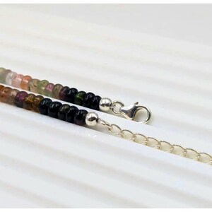 May include: A multi-colored gemstone beaded necklace with a silver clasp and chain extender. The beads are round and feature shades of pink, green, black, and brown.