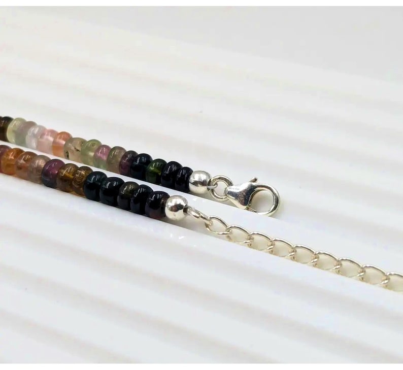May include: A multi-colored gemstone beaded necklace with a silver clasp and chain extender. The beads are round and feature shades of pink, green, black, and brown.