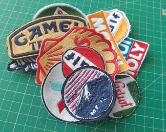 May include: A collection of embroidered patches featuring various logos and designs, including "Camel", "Mobil", "Holy", "Castrol", and a mountain scene. The patches are colorful and have a vintage aesthetic.