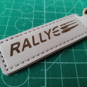 Pode incluir: Um porta-chaves retangular de couro cinza claro com a palavra "RALLYE" impressa em castanho. O porta-chaves tem um anel de metal e uma borda costurada. O fundo é uma base de corte verde.