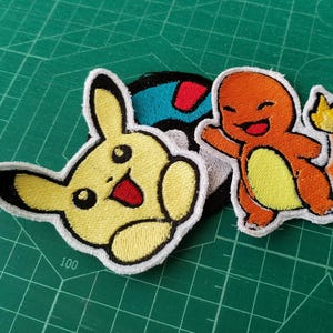 Puede incluir: Dos parches bordados para planchar con personajes de Pokemon. El primer parche es un Pikachu amarillo con una sonrisa roja. El segundo parche es un Charmander naranja con una llama de cola amarilla.