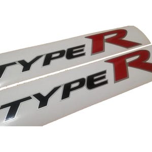Op de afbeelding: Twee witte stickers met de tekst "TYPE R" in zwarte en rode letters. De "R" is rood met een zilveren rand en de "TYPE" is zwart met een zilveren rand.
