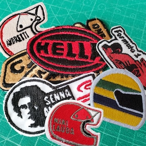 Pode incluir: Uma coleção de patches bordados com temas de corrida. Os designs incluem capacetes, logotipos e os nomes "Andretti", "Senna" e "Niki Lauda". Os patches são em várias cores, incluindo vermelho, preto, branco e amarelo, com costuras detalhadas.