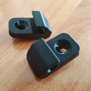 May include: Two small, black plastic clips with a rectangular shape and a curved hook. Each clip has a circular hole. The clips are on a light brown wooden surface.