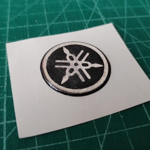May include: A round, black and silver Yamaha logo sticker on a white square. The Yamaha emblem features a stylized design with three outward-pointing arrows. The sticker is on a green cutting mat.