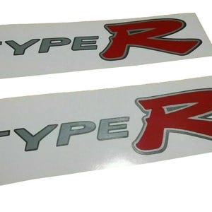 Könnte beinhalten: Zwei weiße Aufkleber mit dem Text "TYPE R" in silberner und roter Schrift. Das "R" ist stilisiert und größer als das Wort "TYPE".