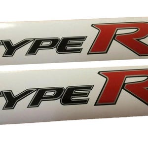 Op de afbeelding: Twee witte stickers met zwarte en rode letters. De stickers zeggen "TYPE R" in een vetgedrukt, gestileerd lettertype. De "R" is rood en de "TYPE" is zwart.