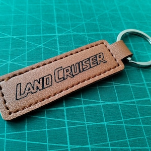 Könnte beinhalten: Ein brauner Leder-Schlüsselanhänger mit dem schwarzen Schriftzug "LAND CRUISER". Der Schlüsselanhänger hat ein rechteckiges Etikett mit genähten Kanten und einen silbernen Metallring. Der Hintergrund ist eine grüne Schneidematte.