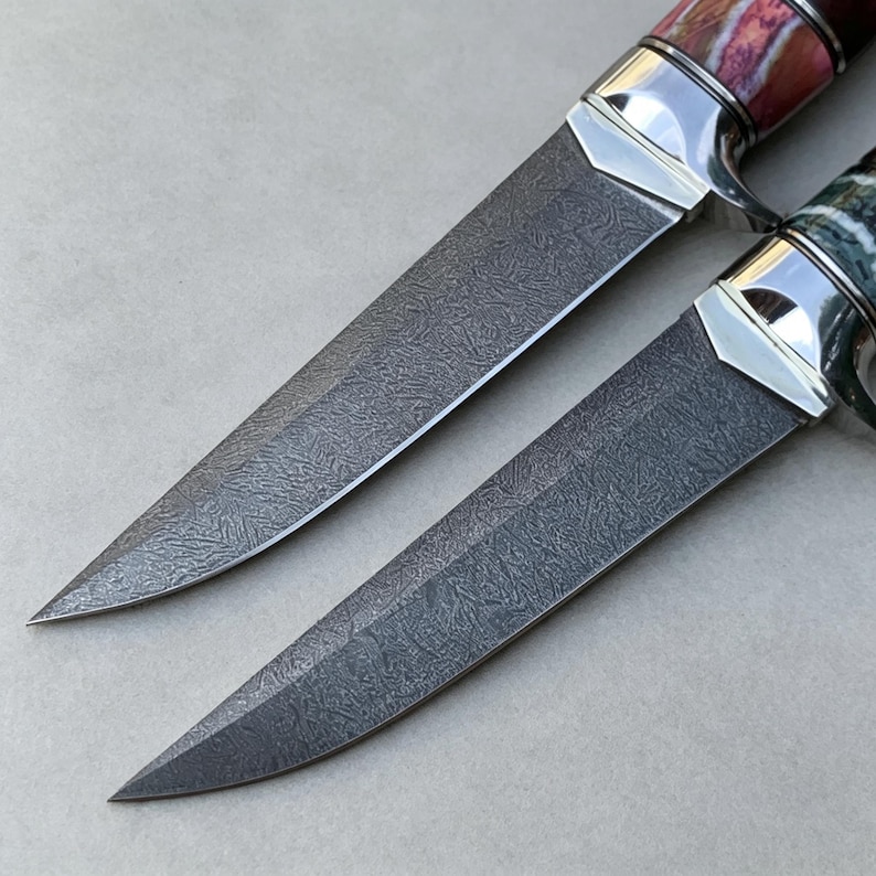 May include: Two knives with dark grey patterned blades featuring a complex, swirling texture. Each knife has a polished silver metal bolster and a handle with visible colorful, marbled material. The blades are pointed and sharp in appearance.