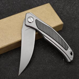 May include: A partially open folding knife with a silver blade and handle. The handle has a carbon fibre inlay and a silver pivot. The knife is resting on a brown cardboard box.