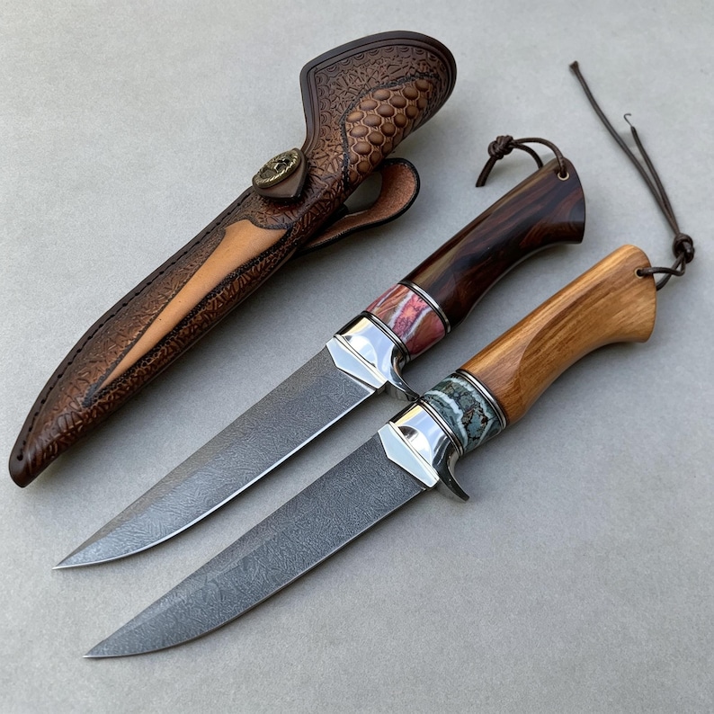 May include: Two fixed-blade knives with Damascus steel blades and polished metal bolsters. One handle is dark wood with a pinkish stone inlay, the other is light wood with a turquoise stone inlay. A brown leather sheath with embossed patterns is included.
