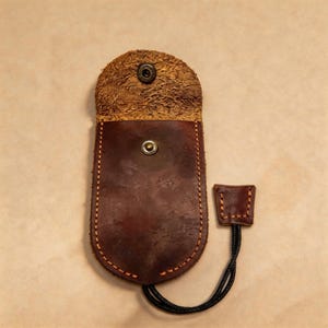 Könnte beinhalten: Eine dunkelbraune Ledertasche mit abgerundeter Klappe und einer strukturierten, hellbraunen Wildlederunterseite. Die Tasche verfügt über einen metallischen Druckknopfverschluss, orangefarbene Kontrastnähte an den Kanten und eine schwarze Kordel.