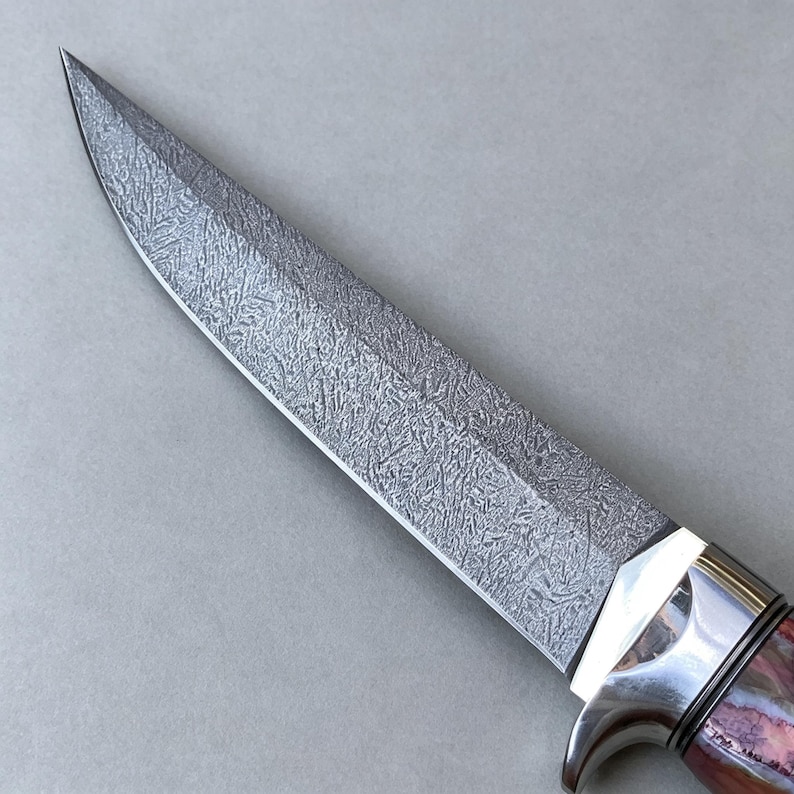 May include: A fixed-blade knife featuring a pointed steel blade with a distinct, intricate, etched crystalline pattern across its surface. The blade connects to a polished silver-colored metal bolster and a handle with reddish-brown tones.