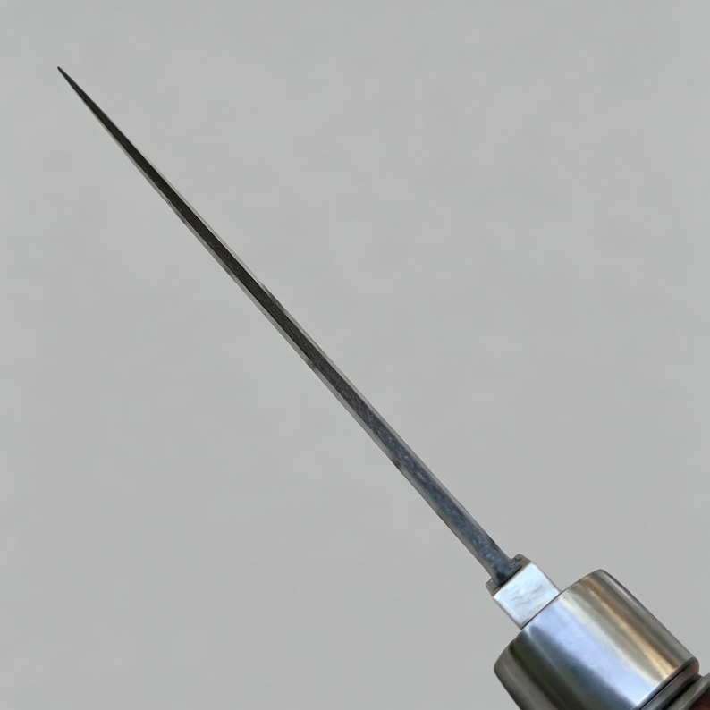 May include: A sharp, thin, metallic blade extending from a cylindrical handle. The blade is silver-toned with a pointed tip and a straight edge, showing a visible transition point where it connects to the smooth, polished metal bolster of the handle.