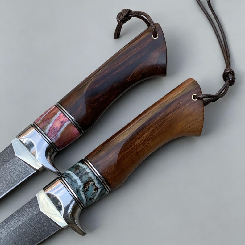 May include: Two knives with dark wood handles, polished metal bolsters, and leather lanyards. One handle features a pink and white stone inlay, while the other has a blue-green and white stone inlay. The blades have a visible dark patterned finish.