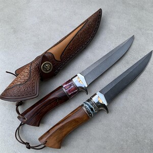 May include: Two fixed-blade knives with dark grey steel blades and polished metal bolsters. The handles are crafted from polished wood with decorative accents. One knife features a reddish-brown handle, while the other has a lighter brown handle. A brown leather sheath with intricate embossed patterns and a circular metal button is included.