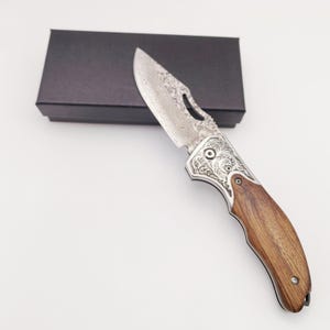 Peut inclure: Un couteau pliant argenté avec un manche en bois et des motifs complexes sur la lame. Le couteau est fermé et repose sur une boîte noire.