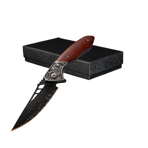 Pode incluir: Um canivete dobrável com uma lâmina escura estampada e um cabo de madeira castanho-avermelhada com um reforço de metal prateado com gravações intrincadas, apresentado ao lado de uma caixa de presente retangular preta texturizada.