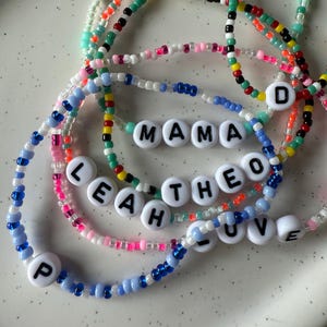 Könnte beinhalten: Eine Sammlung farbenfroher Perlenarmbänder. Einige Armbänder haben weiße Buchstabenperlen, die Namen und Wörter wie "MAMA", "LEAH", "THEO", "LOVE" und den Buchstaben "D" buchstabieren. Die Armbänder sind in verschiedenen Farben erhältlich, darunter Blau, Rosa, Grün und Rot.