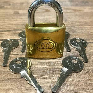 Puede incluir: Un candado de latón con un grillete plateado y varias llaves plateadas dispuestas alrededor. La palabra "HARDENED" está grabada en el grillete. El candado tiene un logotipo circular.