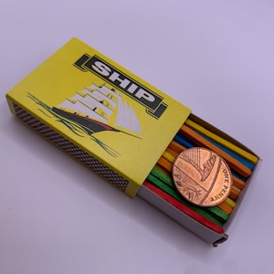 Op de afbeelding: Een gele luciferdoos met een scheepsafbeelding en het woord "SHIP" erop gedrukt. De doos is open en bevat een stapel kleurrijke houten lucifers en een koperen cent.