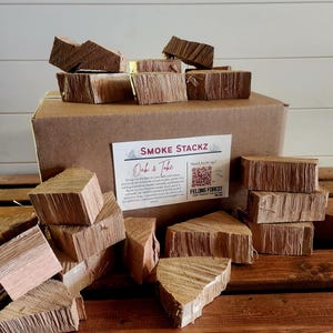 May include: A collection of oak wood chunks for smoking, displayed on a wooden surface. The chunks are various sizes and shapes, with visible wood grain. A cardboard box and a label with the text "SMOKE STACKZ Oak & Toke" are also present.
