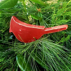 May include: A vibrant red wooden bird ornament with white accents, nestled among lush green pine needles and leaves. The bird is a decorative element, possibly for holiday or seasonal decor.