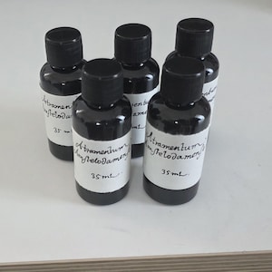 May include: Five small, dark bottles with black caps are arranged on a white surface. Each bottle has a white label with handwritten text, including "Atramentum myfelodamen" and "35 mL". The bottles are filled with a dark liquid.