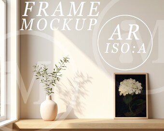 Op de afbeelding: Een wit ingelijst beeld met een witte hortensia op een zwarte achtergrond. Het beeld staat op een licht houten plank met een vaas met groene bladeren en een witte vaas.